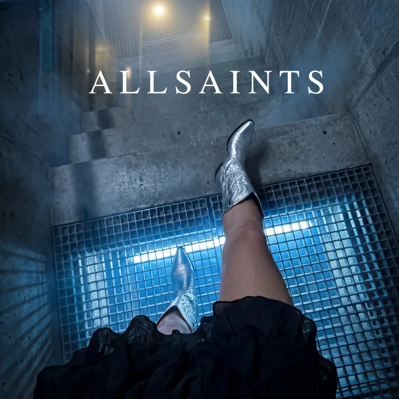 AllSaints key visual: silver cowboy boots on a lit grate in a concrete stairwell, with the AllSaints name